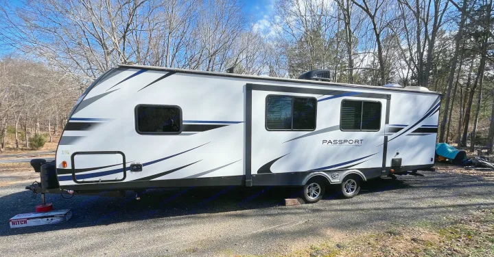 White Passport travel trailer RV rental parked on gravel, featuring dual axles, blue accents, and dark windows, set against bare trees.. Keystone RV Passport Grand Touring 2019
