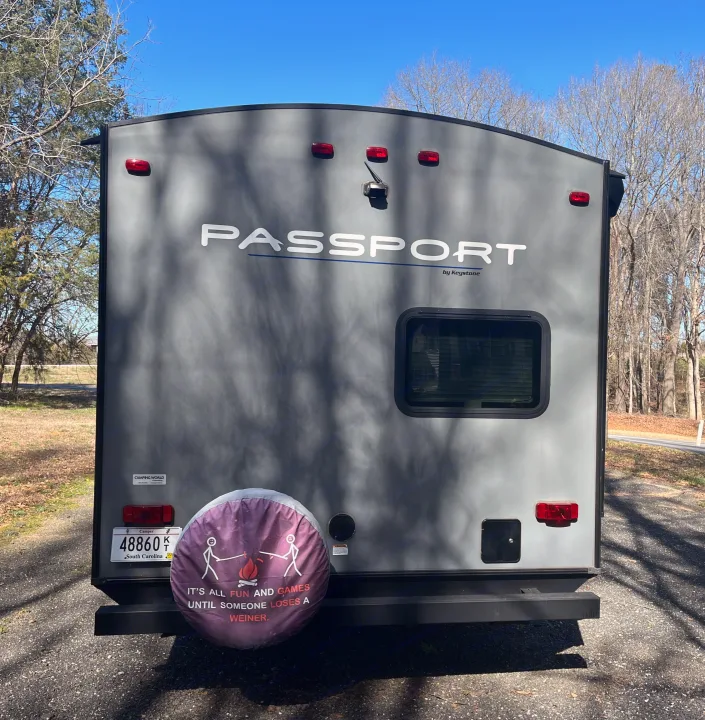 Rear view of a gray Passport travel trailer, an ideal rv rental, with a humorous camping spare tire cover and SC plate.. Keystone RV Passport Grand Touring 2019