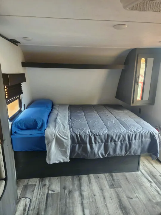 Cozy RV rental bedroom featuring a full bed with blue sheets & grey comforter, overhead storage, window, and wood-look flooring.. Forest River ASPEN TRAIL 2024