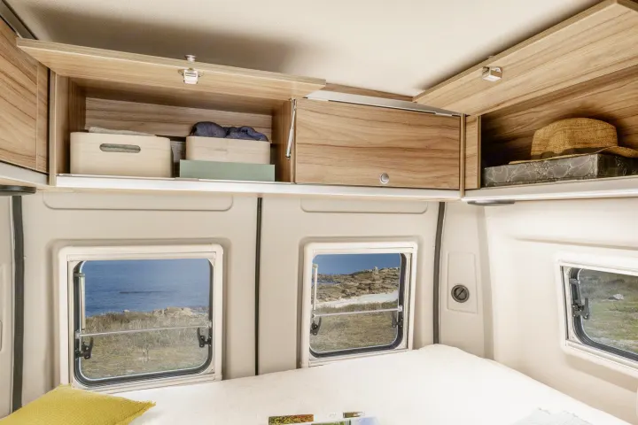 RV rental: Cozy bedroom interior with open wood cabinets showing storage boxes & a hat, alongside scenic coastal views.. Hymer Ayers Rock 2024