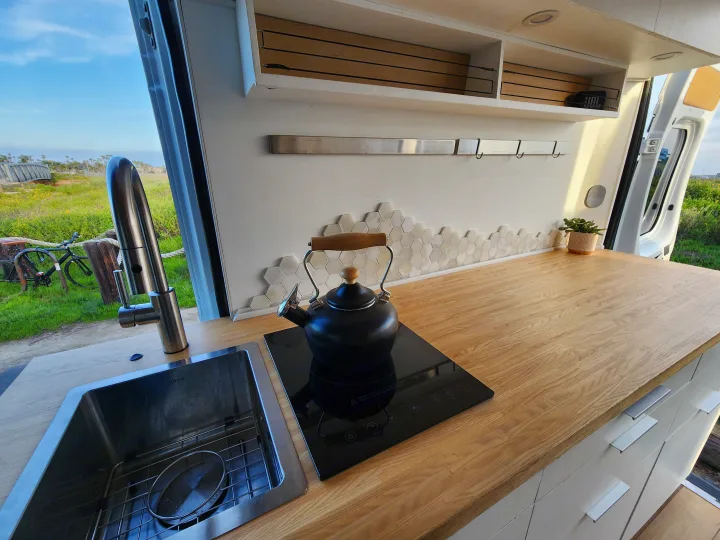 Stylish RV rental kitchen with sink, induction cooktop, and wood counter. Green landscape outside.. Dodge Sprinter Van 2015