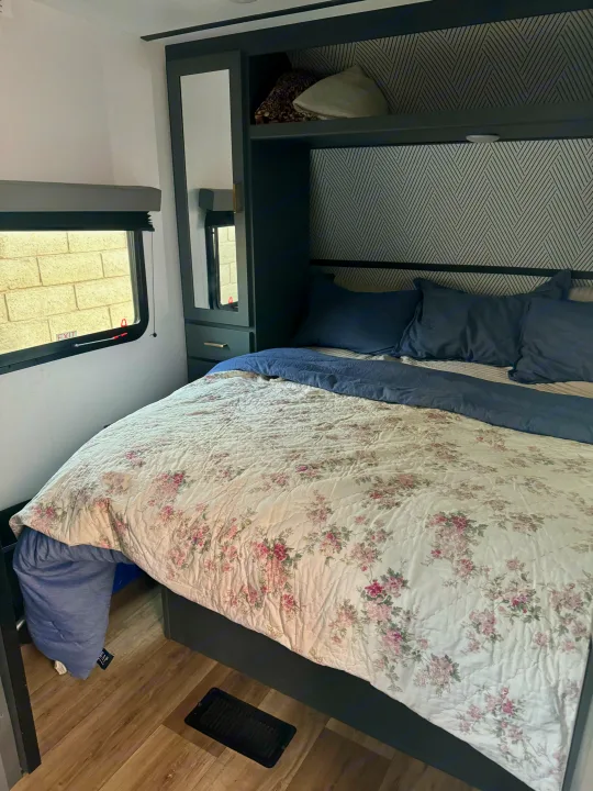 RV rental bedroom: floral bed, blue pillows, dark built-in storage with mirror, window, and patterned wall.. Keystone RV Bullet Crossfire 2022