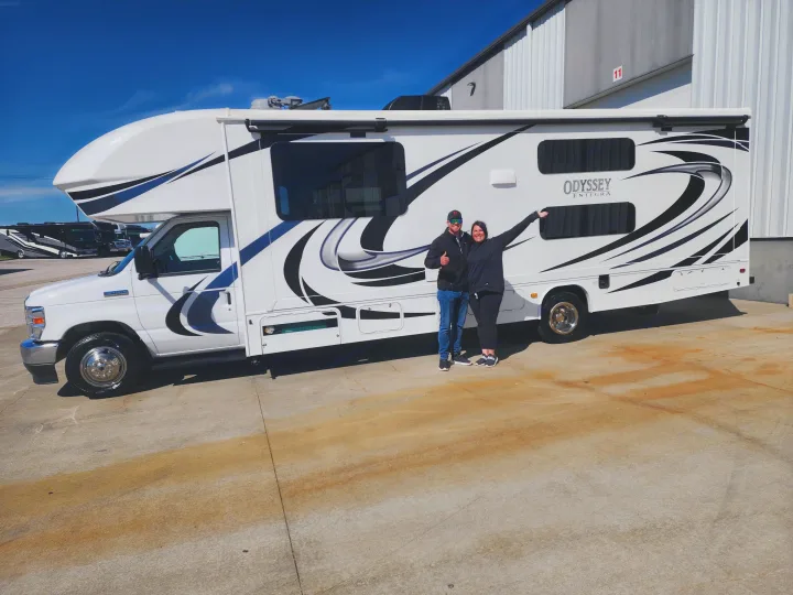 A happy couple poses beside their white recreational vehicle, an Odyssey Entegra RV rental, under a clear blue sky.. Entegra Coach Odyssey 2021