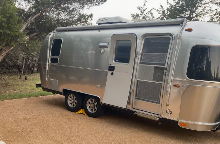 Shiny silver Airstream travel trailer, a perfect RV rental for camping adventures. Parked on gravel with the door open at a wooded site.. Airstream Flying Cloud 2018