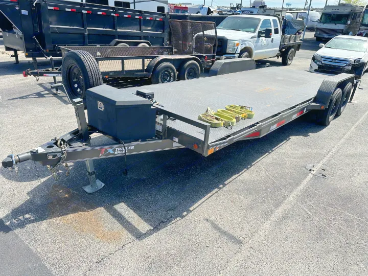 A black car hauler trailer with tie-downs, parked in a lot. An RV rental, pickup truck, and other trailers are visible in the background.. East Texas Trailers Car Hauler 2021