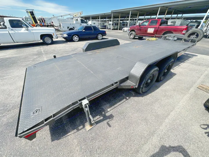 Black car hauler parked on asphalt. A red RV rental truck, other vehicles, and RVs are visible under covered parking.. East Texas Trailers Car Hauler 2021