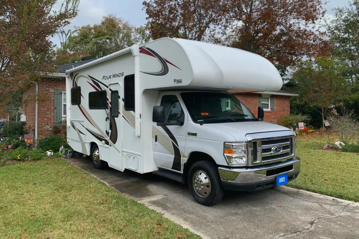 This white Four Winds Class C RV rental is parked on a driveway beside a brick house and fall foliage.. Thor Four Winds 2020