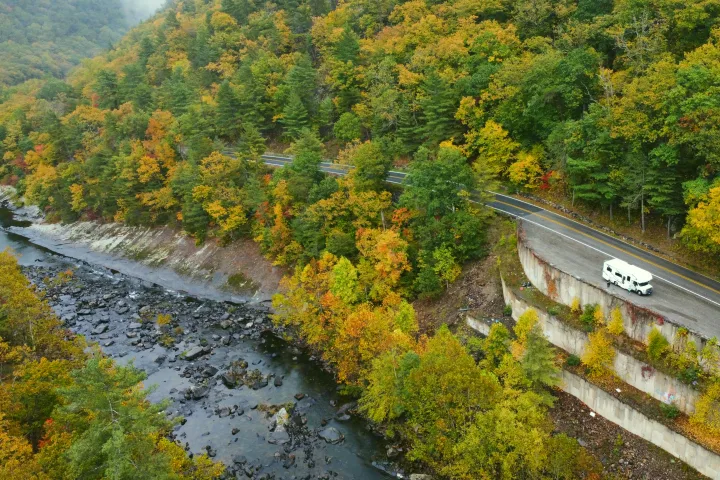Scenic aerial view: RV rental parked on a mountain road winding above a rocky river, surrounded by vibrant autumn forest.. Thor Four Winds 2020