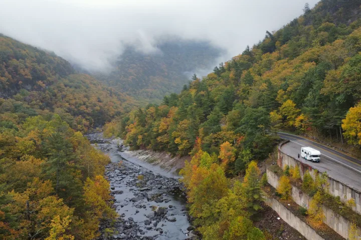 An RV rental parked on a scenic mountain road, overlooking a river valley with vibrant fall foliage and fog.. Thor Four Winds 2020