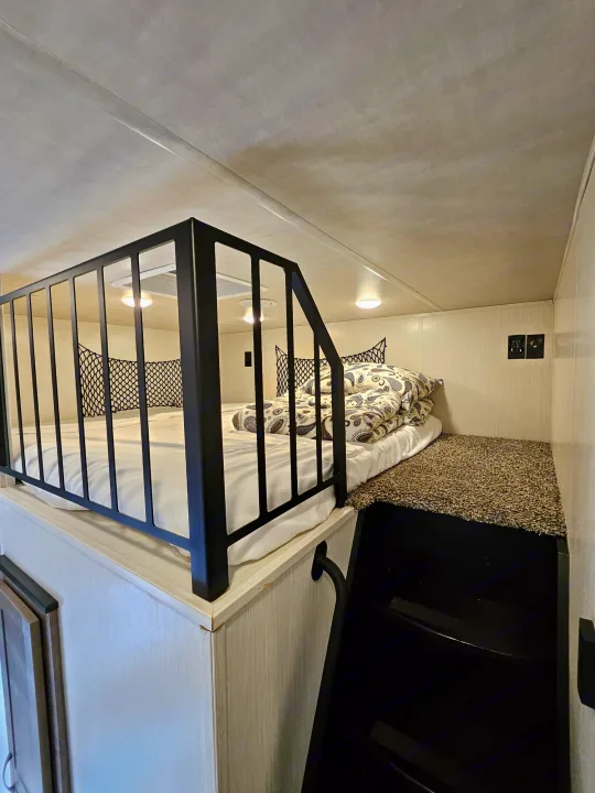 An RV rental's inviting loft bed, complete with a dark safety railing, patterned bedding, and carpeted floor leading to black stairs below.. Forest River Cedar Creek Fifth Wheel 2022