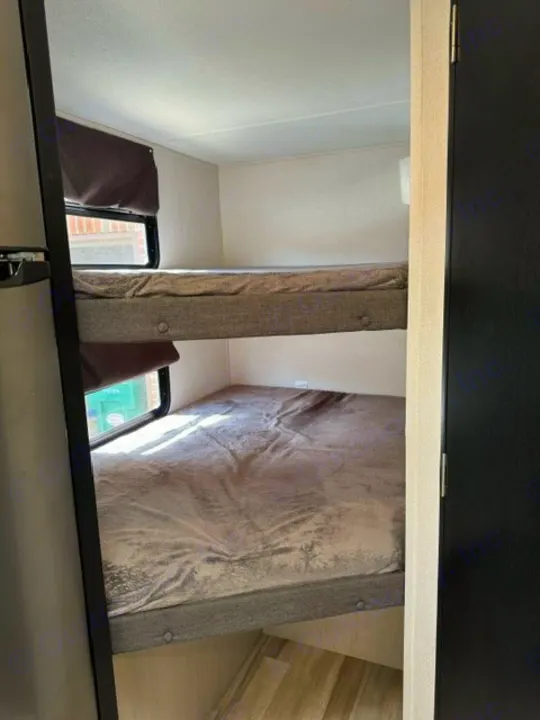 An RV rental's compact bunk bed room features two grey-covered beds, one above the other, with windows on the left.. Forest River Tracer 2022