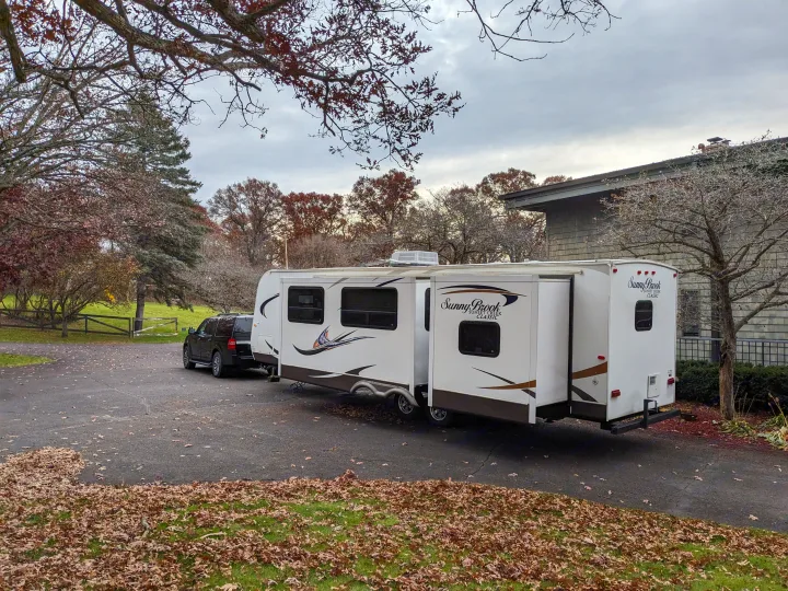 White travel trailer RV rental, with extended slide-out, parked on a driveway beside a black SUV. Autumn leaves visible on the ground.. Winnebago Sunset Creek 2014