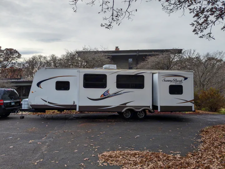 A white travel trailer RV rental, Sunny Brook Sunset Creek Classic, is parked with its slide-out extended on an asphalt driveway.. Winnebago Sunset Creek 2014