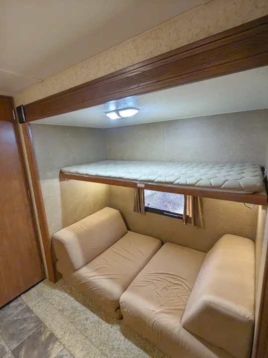 Compact RV rental interior featuring a top bunk bed with mattress, lower convertible sofa, and an emergency exit window.. Winnebago Sunset Creek 2014