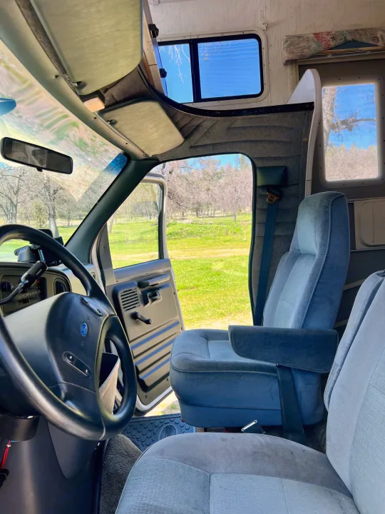 Inside an RV rental: driver's view of the steering wheel, blue seats, and bunk. Door open to a sunny, green field.. Ford Econoline 1994