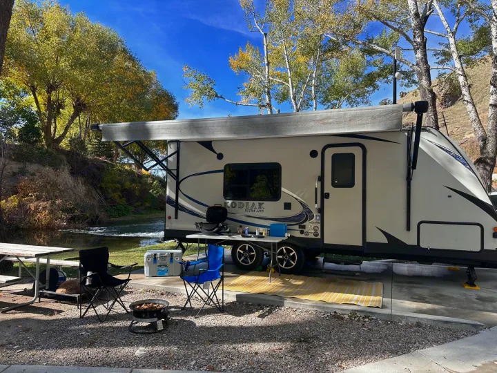A Kodiak rv rental is perfectly set up at a scenic riverside campsite, featuring a picnic table, chairs, & fire pit for comfort.. Dutchmen Kodiak Ultimate 2018