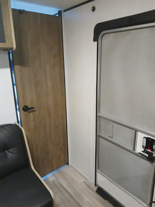 Inside an RV rental, a wooden interior door stands open next to a light wall and the main screen entrance door, showing flooring.. Heartland RVs Pioneer 2022