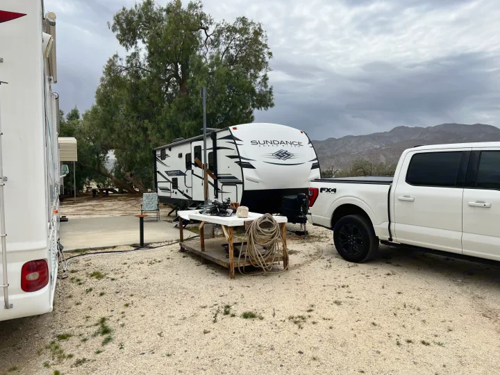 An RV rental travel trailer and pickup truck parked at a gravel campsite with trees and distant mountains under a cloudy sky.. Heartland RVs Sundance Ultra Lite 2022