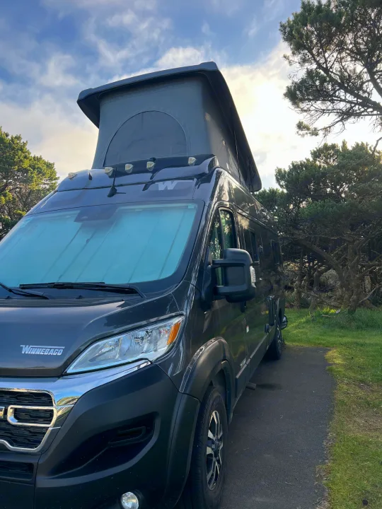 Dark grey camper van with pop-top roof parked in a natural setting with gnarled trees, perfect for an rv rental adventure.. Winnebago Solis 2023