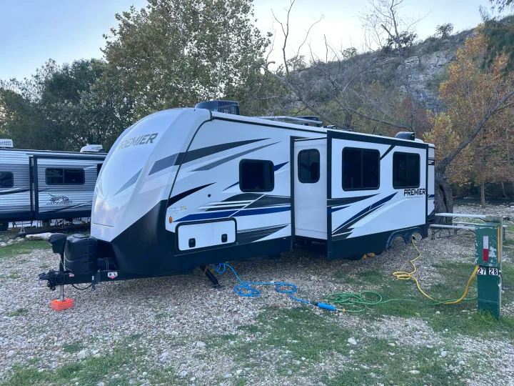 Experience nature! This Premier travel trailer RV rental is set up at a gravel campsite with utility hookups and trees.. Keystone RV Bullet Premier Ultra Lite 2022