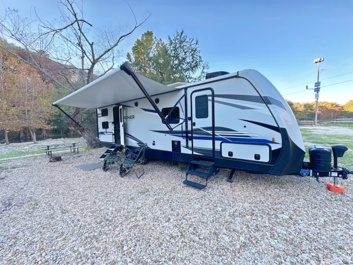 This white RV rental travel trailer with an extended awning is parked at a scenic gravel campsite. Perfect for outdoor getaways.. Keystone RV Bullet Premier Ultra Lite 2022