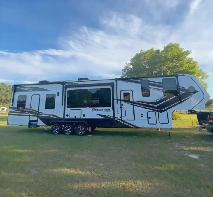 Explore the outdoors with this large, striking white and black fifth-wheel RV rental. Parked in a sunny green field.. Grand Design Momentum M-Class Toy Hauler 2023