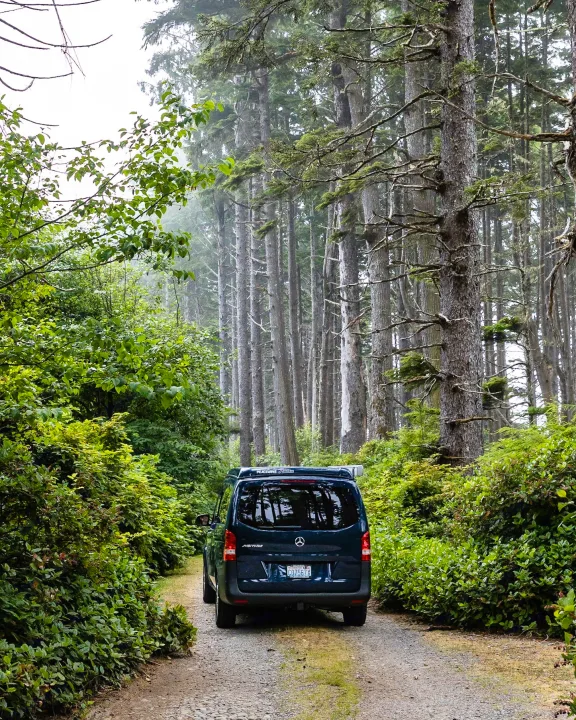 Dark blue Peacevans Mercedes-Benz RV rental on a gravel road, amidst tall, misty evergreen trees. Ideal for a nature escape.. Mercedes-Benz metris 2022