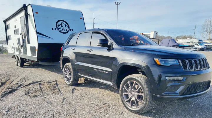 Black Jeep Grand Cherokee towing a white Ozark travel trailer on a gravel lot. Ideal for an RV rental getaway.. Forest River ozark 2440bhkx 2024