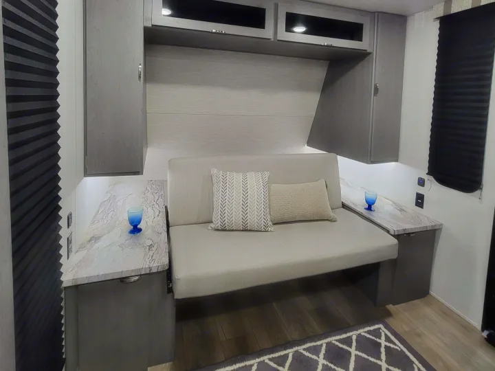 Modern RV rental interior with a light sofa/bed, side tables holding blue glasses, overhead cabinets, and dark pleated blinds.. Forest River ozark 2440bhkx 2024
