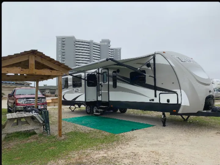 RV rental: A travel trailer with awning extended, parked beside a truck, picnic table, and wooden shelter. Buildings are in the distance.. Keystone Laredo 2015