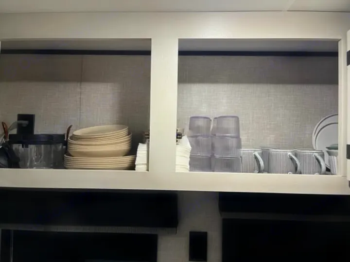 RV rental kitchen: Stacked plates, clear cups, striped mugs, and a food processor fill an open cabinet.. Highland Ridge RV Open Range 2023
