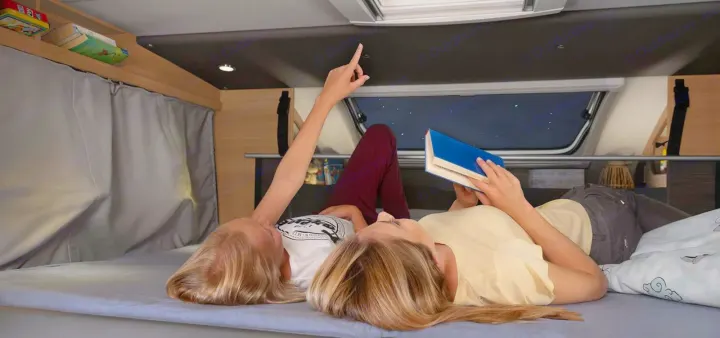 Experience starry nights in an RV rental. Two girls relax in a comfy RV bed, one stargazing, one reading.. Knaus live wave 2024