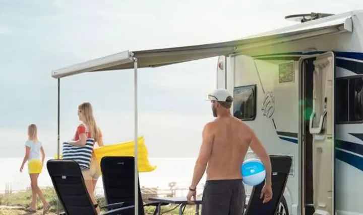Relaxing RV rental vacation by the water. An awning shades a family, chairs, and beach gear next to a white RV.. Knaus live wave 2024