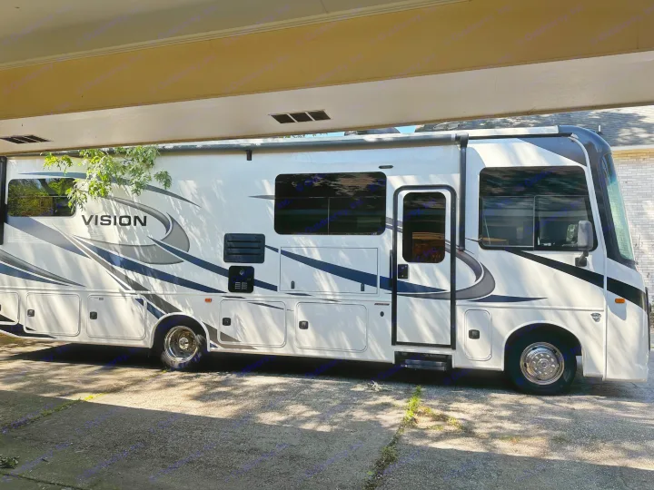 A large white and black RV, labeled 'VISION', is parked under a covered structure with dappled sunlight. Perfect for an rv rental experience.. Entegra Coach Vision 2021