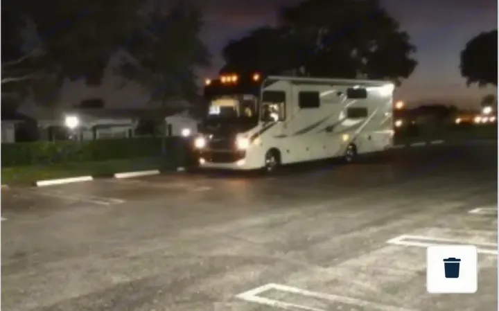 Night scene: a sleek white RV rental with its lights on, parked in a paved lot, with trees and distant lights visible.. Entegra Coach Vision 2021