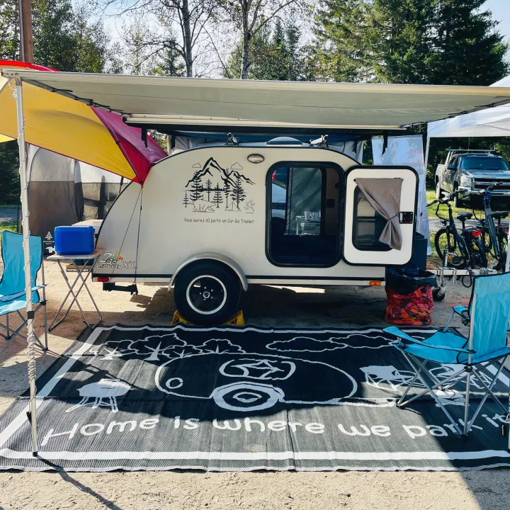 An RV rental camper with an extended awning and camping gear, including chairs and a mat, set up at a scenic campsite.. Car-gotrailers liberty2goXL 2021
