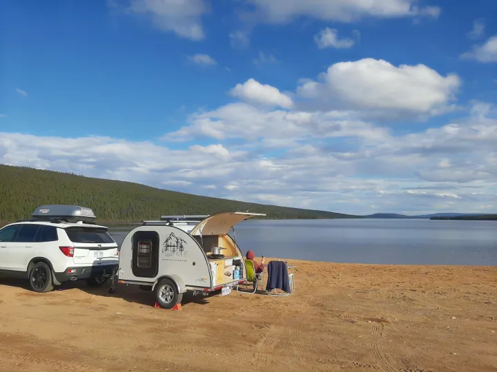 Serene lakeside RV rental scene: a car and teardrop trailer parked for camping, a person relaxing by the water.. Car-gotrailers liberty2goXL 2021