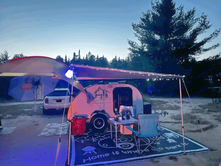A cozy teardrop rv rental setup with a lighted awning at dusk. "Home is Where We Park It" mat adds to the inviting camping scene.. Car-gotrailers liberty2goXL 2021