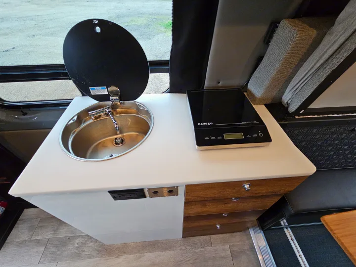 Inside this RV rental, find a compact kitchenette with a sleek induction cooktop, sink, and storage drawers. Perfect for travel.. Storyteller Overland Stealth Mode 2021
