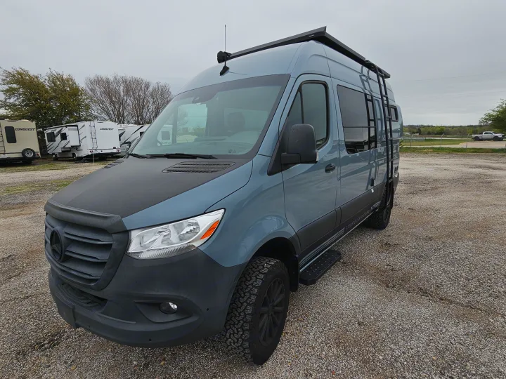 RV rental: A grey Mercedes-Benz Sprinter camper van with a black hood & off-road tires, parked on a gravel lot with other RVs.. Storyteller Overland Stealth Mode 2021