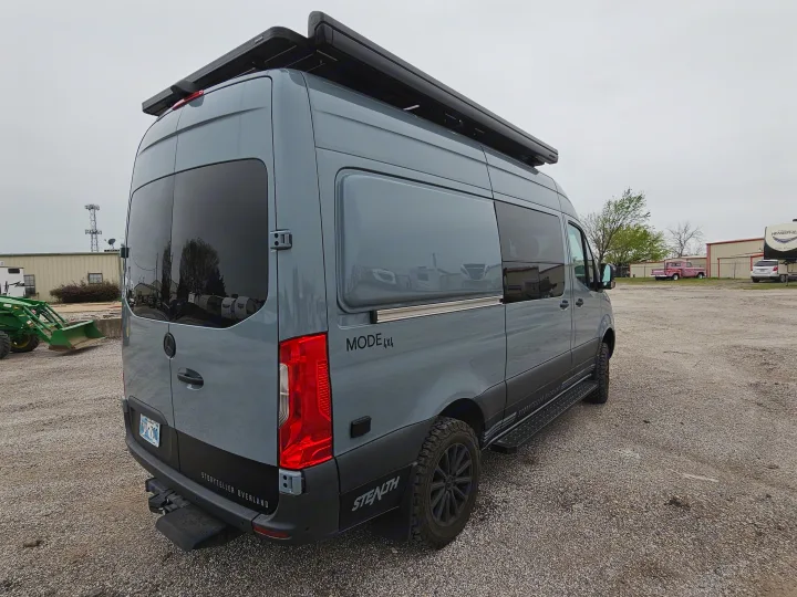 A grey Storyteller Overland 4x4 Sprinter camper van, featuring a roof awning and rugged tires, awaits your next rv rental journey.. Storyteller Overland Stealth Mode 2021