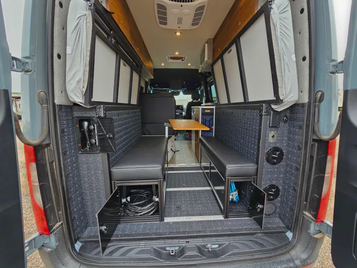 Step into this spacious RV rental camper van interior, featuring rear-facing bench seating, a dining table, overhead cabinets, and practical storage.. Storyteller Overland Stealth Mode 2021