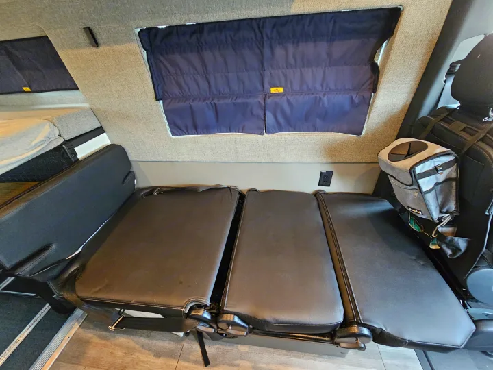 RV rental interior: black convertible seating that transforms into a bed, dark window shades, and a raised sleeping area.. Storyteller Overland Stealth Mode 2021