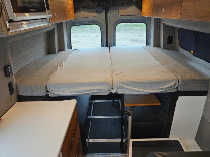 RV rental camper van interior featuring a comfortable convertible bed, microwave, and storage. Rear windows offer a view.. Storyteller Overland Stealth Mode 2021