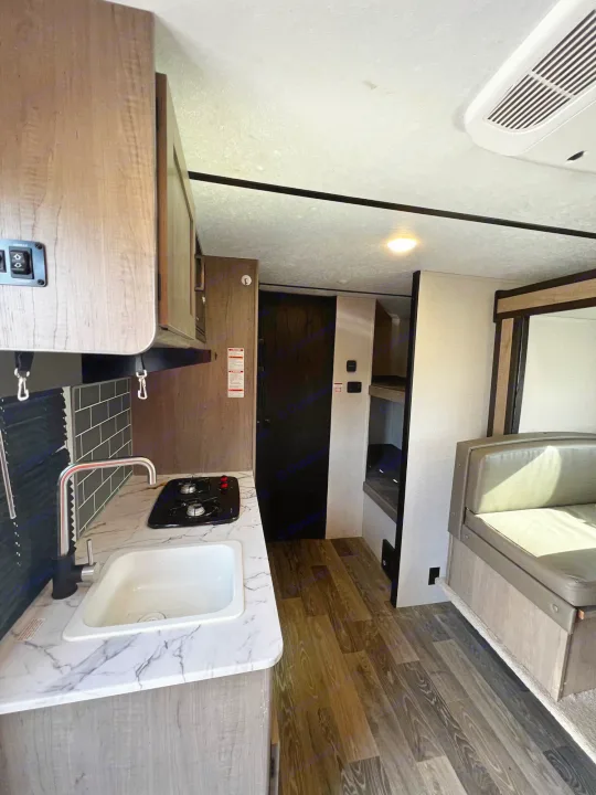 A clean RV rental interior features a kitchen with sink and stovetop, wood-look flooring, and a dinette leading to bunk beds.. Keystone Hideout 2021