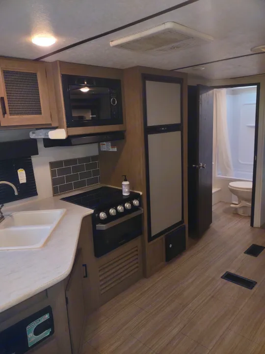 RV rental interior: Kitchen with sink, stove, microwave, and fridge, leading to a bathroom with shower & toilet.. Keystone Hideout 2020
