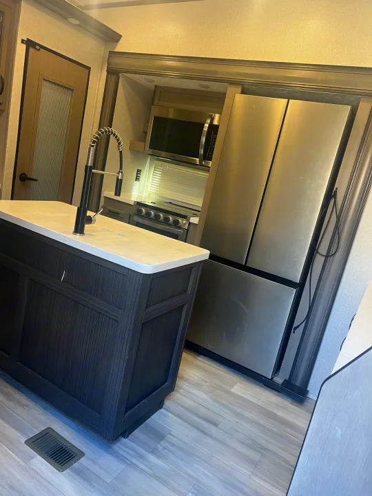 RV rental kitchen: modern island with faucet, stainless steel refrigerator, microwave, stove, and wood-look flooring.. Coachmen Chaparral 2021