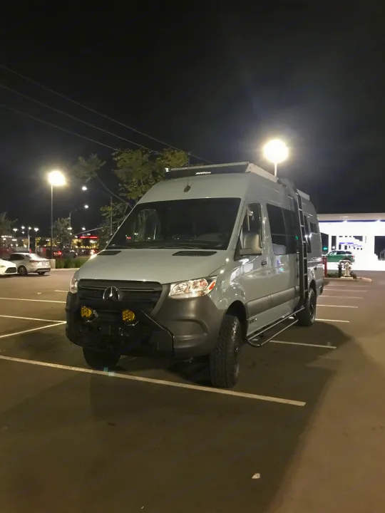 This custom light grey Mercedes-Benz Sprinter adventure van, ideal for RV rental, is parked in a bright nighttime lot.. Mercedes-Benz Sprinter 2022