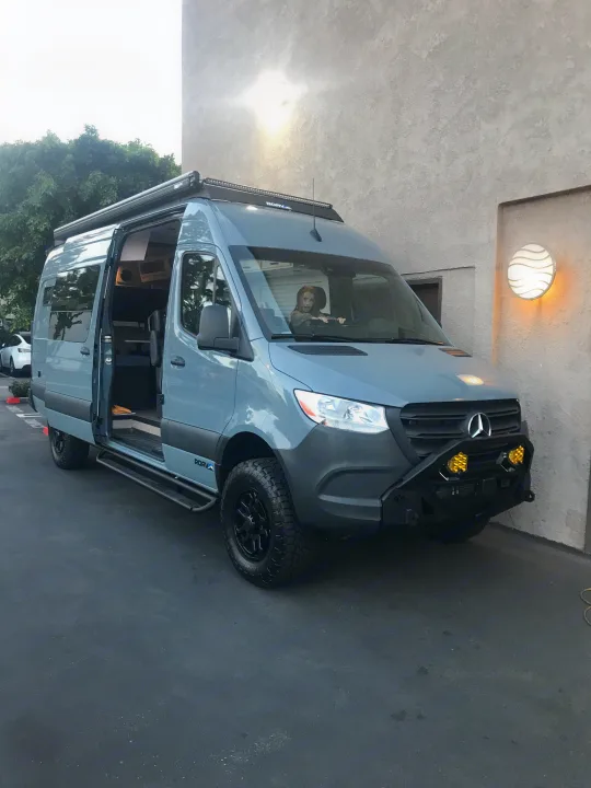 Custom grey Mercedes Sprinter RV rental. Child in driver's seat. Features off-road tires, light bar, and open side door.. Mercedes-Benz Sprinter 2022