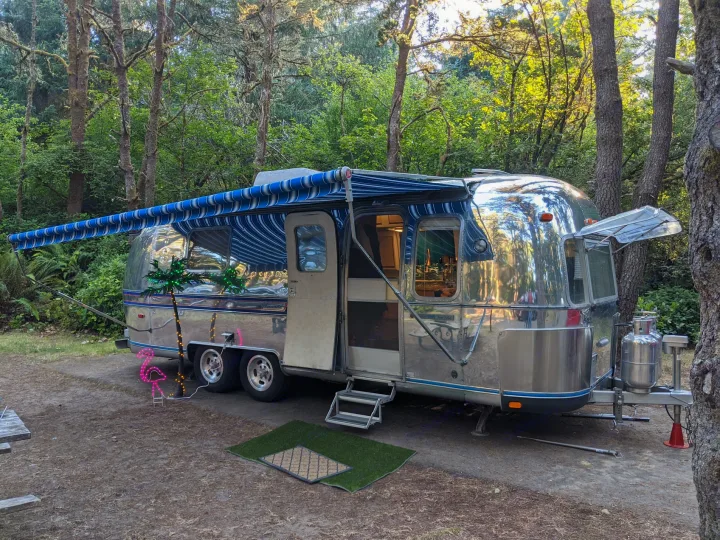 A polished Airstream RV rental, with an extended awning and festive lights, is perfectly set up at a wooded campsite.. Airstream Tradewind 1979